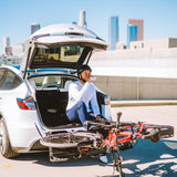 Man relaxing with water bottle next to red electric bike on car hitch rack.